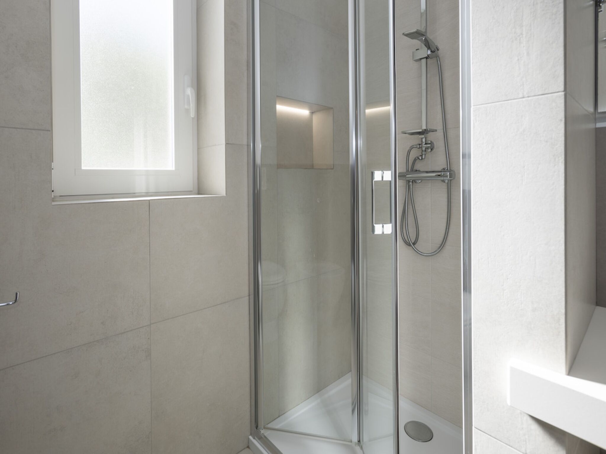 A modern bathroom with a glass-enclosed shower, white tiles, and a window providing natural light.