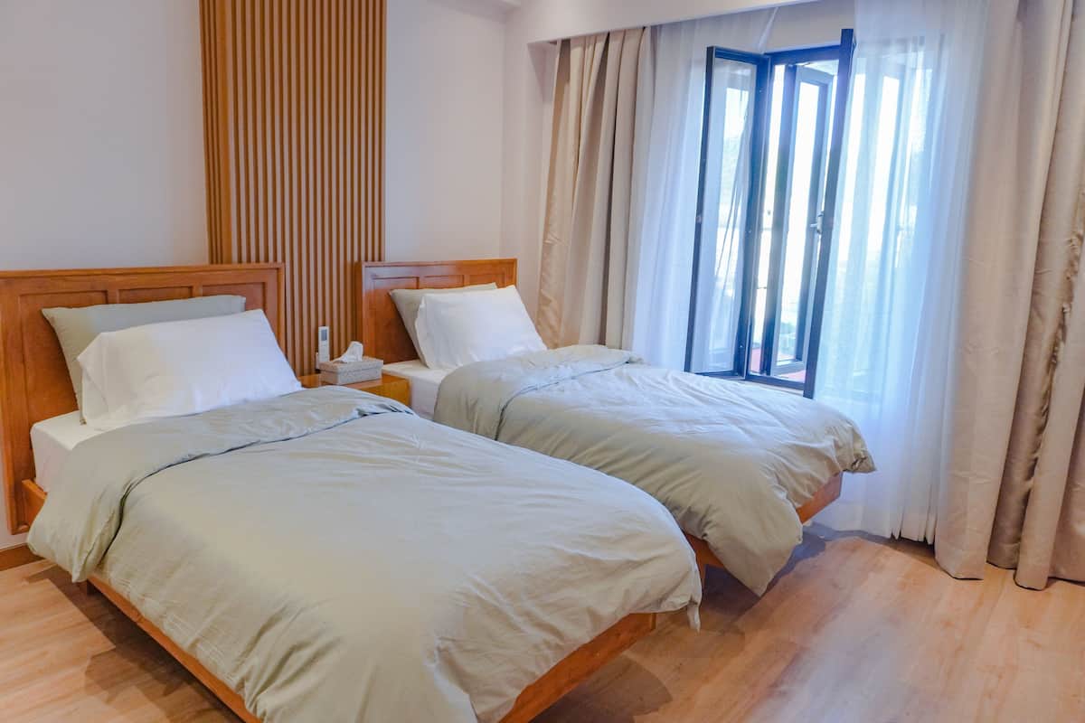 A hotel room with two single beds, a wooden headboard, a bedside table with a lamp, and a window with curtains.