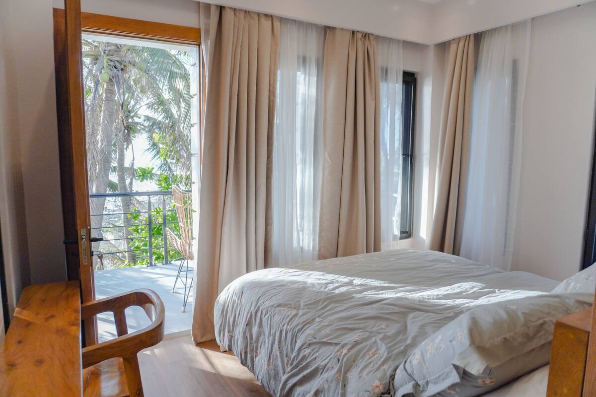 A bedroom with a bed, a wooden desk, and a balcony with curtains.
