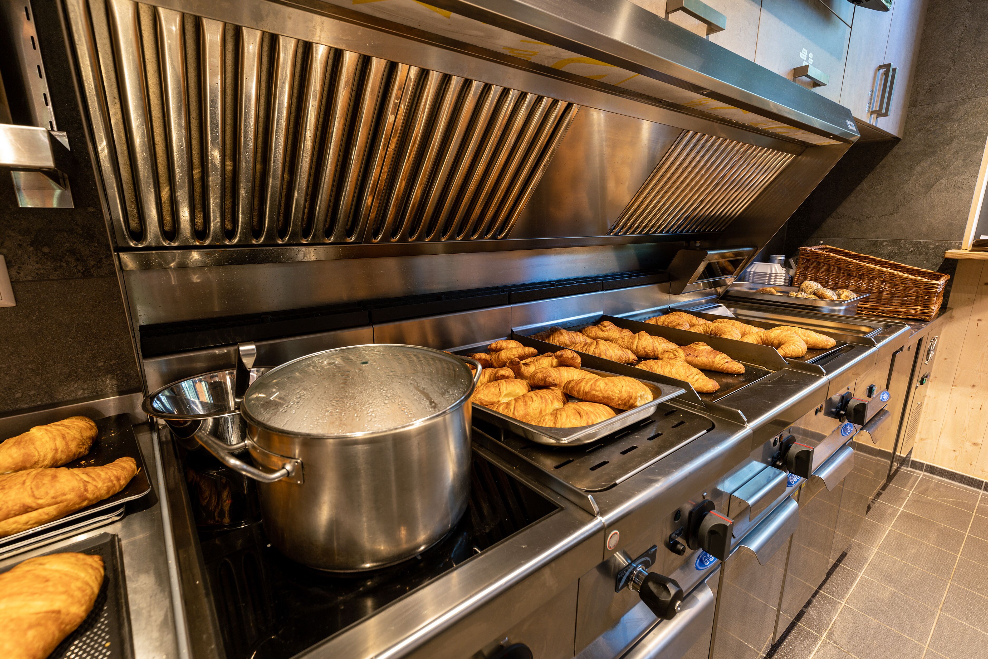 Una cocina comercial con electrodomésticos de acero inoxidable, incluyendo una olla en la estufa y bandejas con productos horneados.