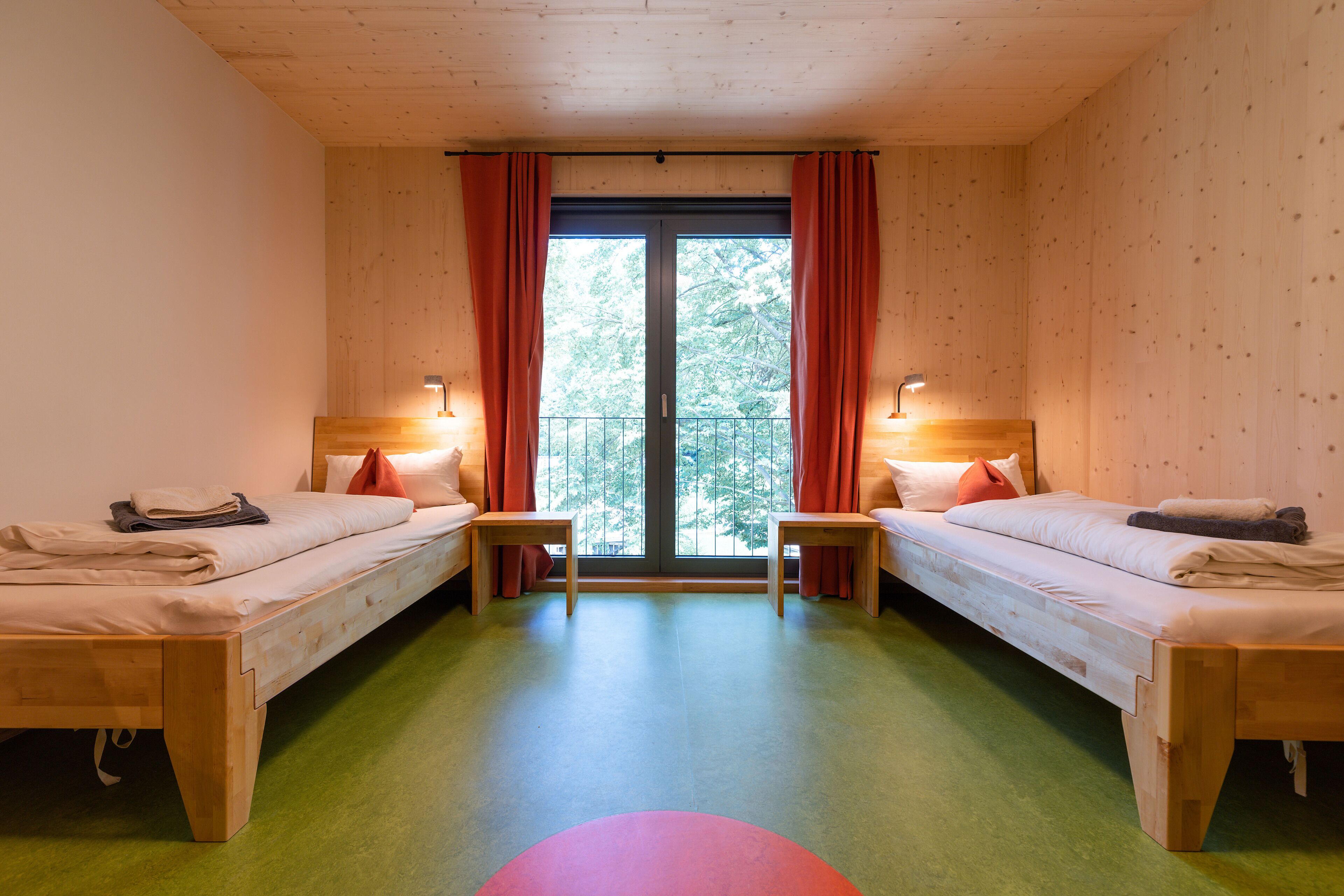Een kamer met twee houten bedden, een groen tapijt en een balkon met rode gordijnen.