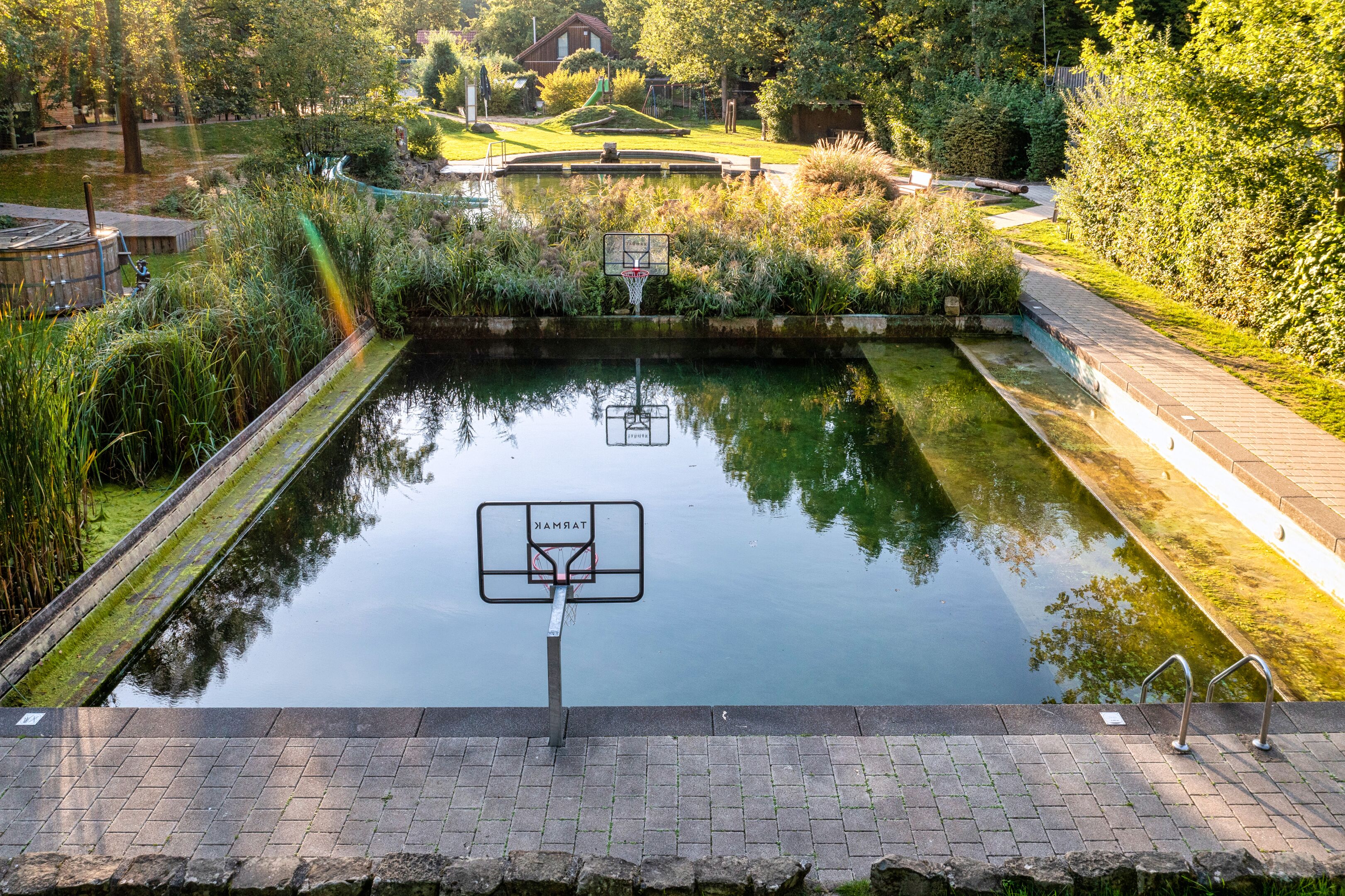 A basketball court with a water feature in the middle, surrounded by greenery and a brick pathway.