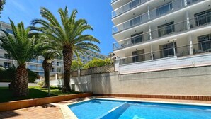 Une piscine bordée de palmiers, un immeuble à plusieurs étages doté de balcons et un ciel d’un bleu limpide.
