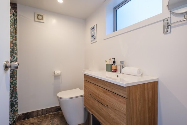A bathroom with a white toilet, wooden vanity, and a window above the sink. - Gulf Island Views - Welcome to Gulf Island Views, a beautifully restored premium holiday cottage with incredible views of the Hauraki Gulf. With two bedrooms and one bathroom this is perfect for a family of four or two couples to share. (Waiheke Island)