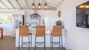 Una cocina moderna con una isla central, tres taburetes de mimbre, iluminación colgante y un ventanal grande.