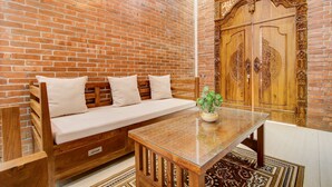 A room with a brick wall, wooden bench with cushions, a glass-top table, and a large wooden door with intricate carvings. - Capital O 93718 Homestay 3 Bidadari (Moyudan)