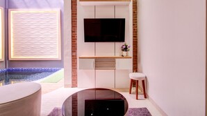 A modern living room with a flat-screen TV mounted on a white wall, a glass coffee table, and a seating area with a cushioned stool. - Capital O 93718 Homestay 3 Bidadari (Moyudan)