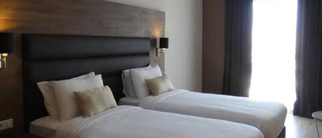 A hotel room with two beds, a dark headboard, bedside lamps, and a large window with curtains.