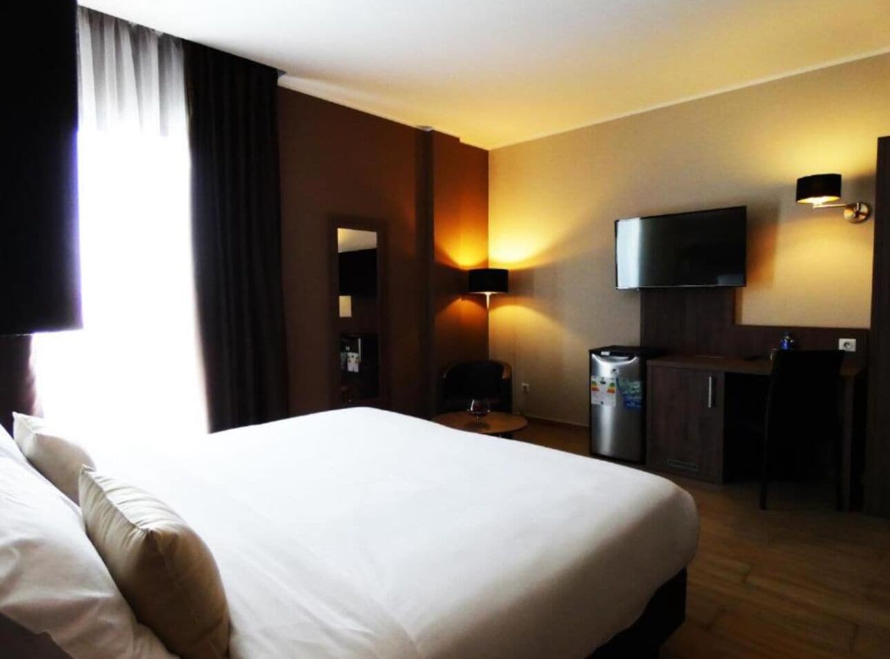 A hotel room with a large bed, a desk, a television, and a minibar.