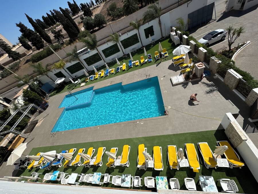 Piscina en la azotea con varios sillones de sol y sombrillas, rodeada de un área ajardinada con árboles y cielo despejado.