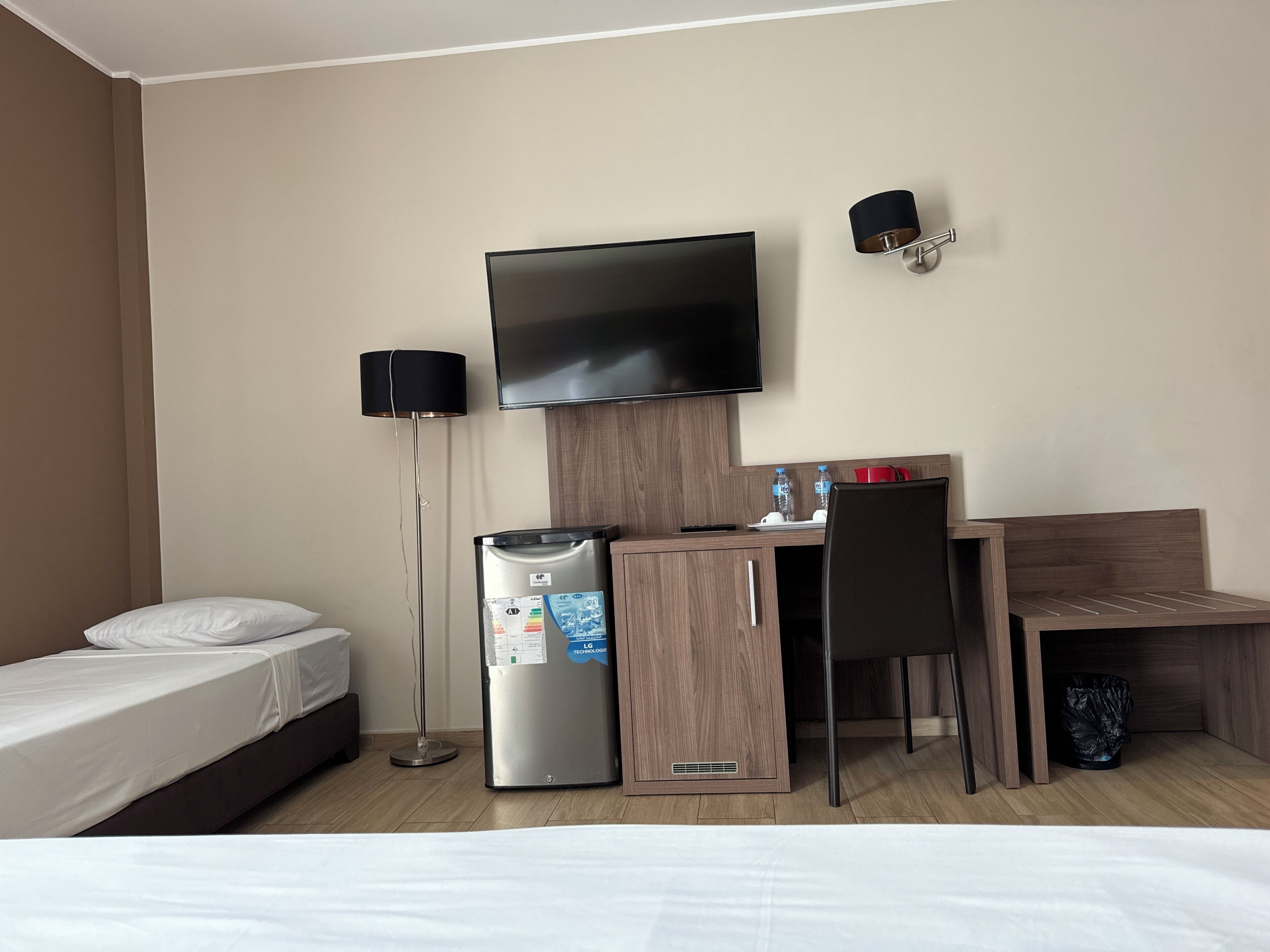 Une chambre d’hôtel comprenant un lit, un bureau avec une chaise, un téléviseur à écran plat, un réfrigérateur, une lampe sur pied et une poubelle.