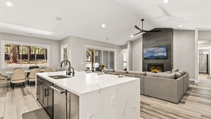 A modern kitchen with a marble island, a dining area with chairs, and a living room with a sofa and fireplace. - Large 5BR w hot tub, fireplace, forest views! (Flagstaff)