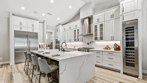A modern kitchen with white cabinetry, a central island with bar stools, stainless steel appliances, and a wine refrigerator. - Large 5BR w hot tub, fireplace, forest views! (Flagstaff)