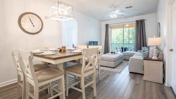 Comedor con mesa y sillas de madera, un reloj en la pared y un balcón con vistas a árboles.