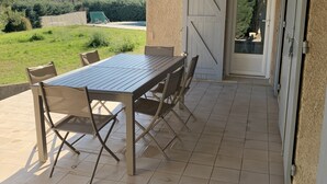 Een overdekt terras met een tafel en stoelen, een schuifpui en uitzicht op de omgeving.