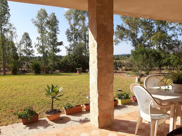 A patio with a table and chairs, surrounded by potted plants and a view of a grassy field with trees. - House immersed in the green, a few steps from the sea (Santa Maria Villaputzu)