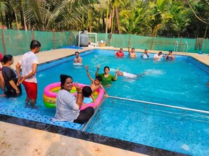 A swimming pool with people enjoying the water, surrounded by a fence and palm trees. - Gokul Cottage and Villa (Alibag)