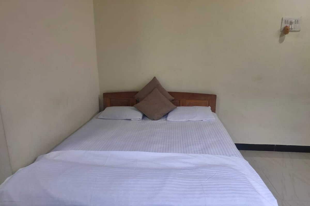 A single bed with a wooden headboard, two pillows, and a white bedspread in a room with plain walls and a visible electrical outlet.