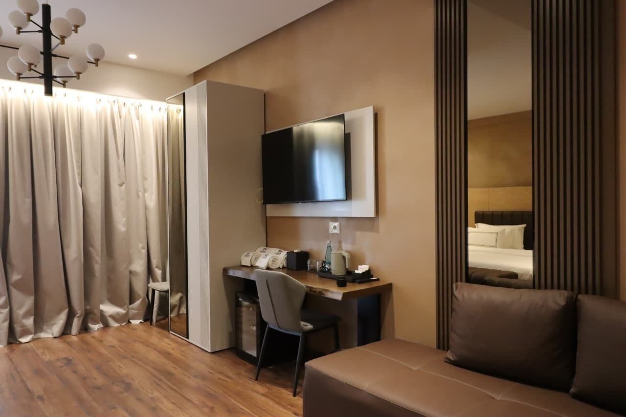 A modern hotel room with a wooden floor, a desk with a chair, a flat-screen TV, a bed with a headboard, and a large mirror.