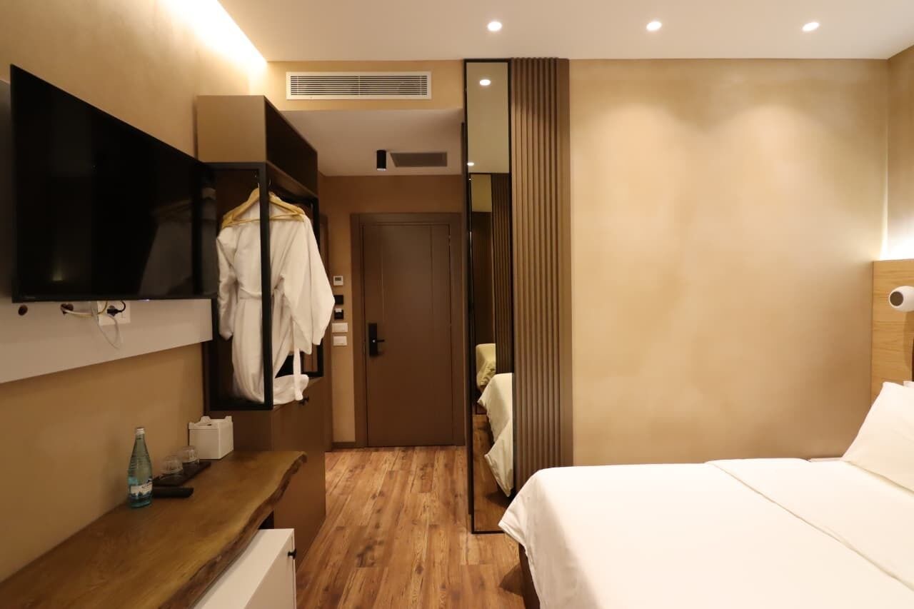 Ein Hotelzimmer mit einem Bett, einem an der Wand montierten Fernseher, einem hölzernen Schminktisch, einem Spiegel und einer Tür zu einem anderen Raum.