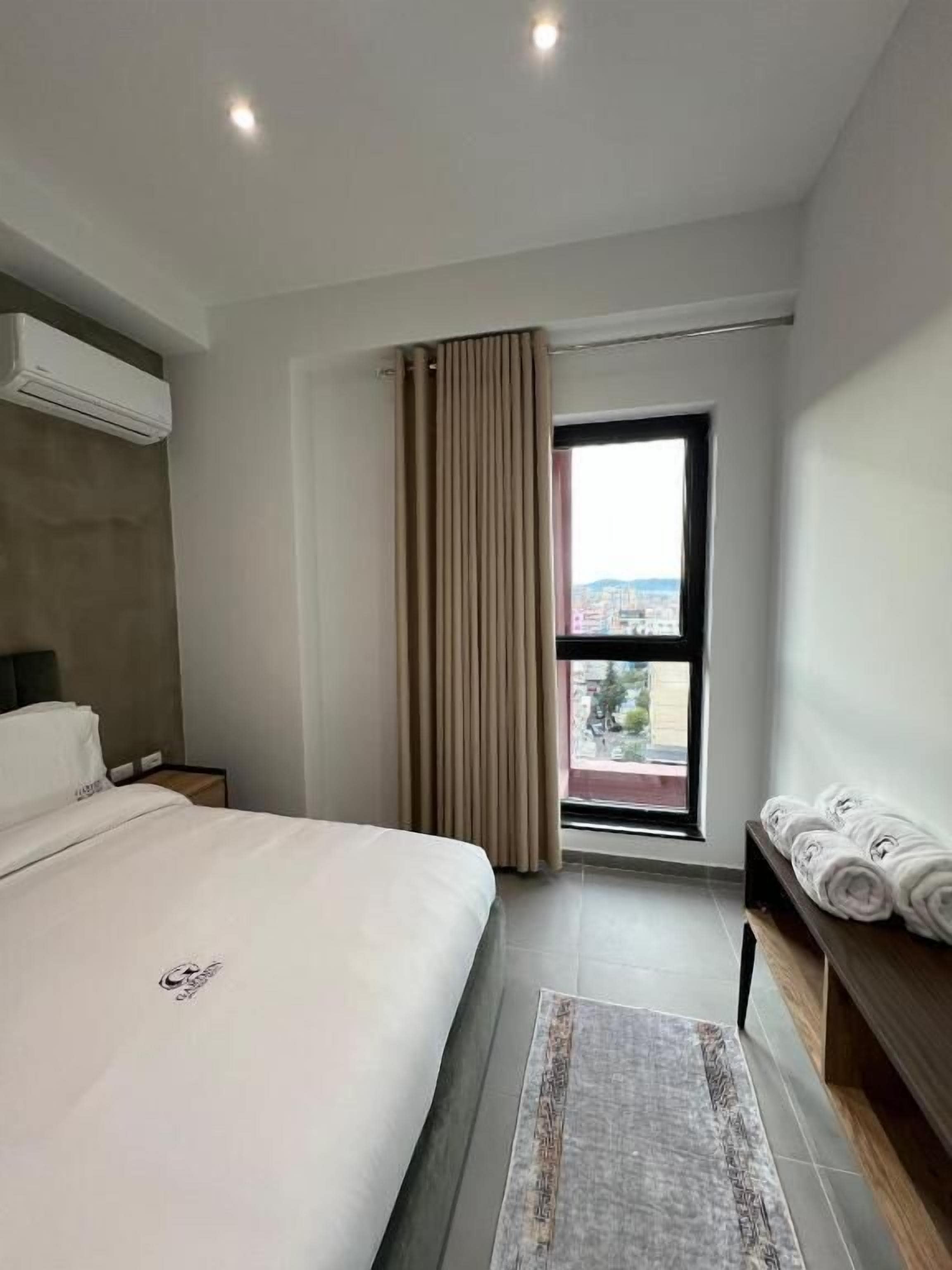 Une chambre d’hôtel moderne avec un grand lit, une table de chevet, une commode, une fenêtre avec des rideaux et une vue sur la ville.