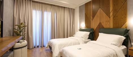 A hotel room with two beds, a wooden desk, a plant, and a view of the outdoors through large curtains.
