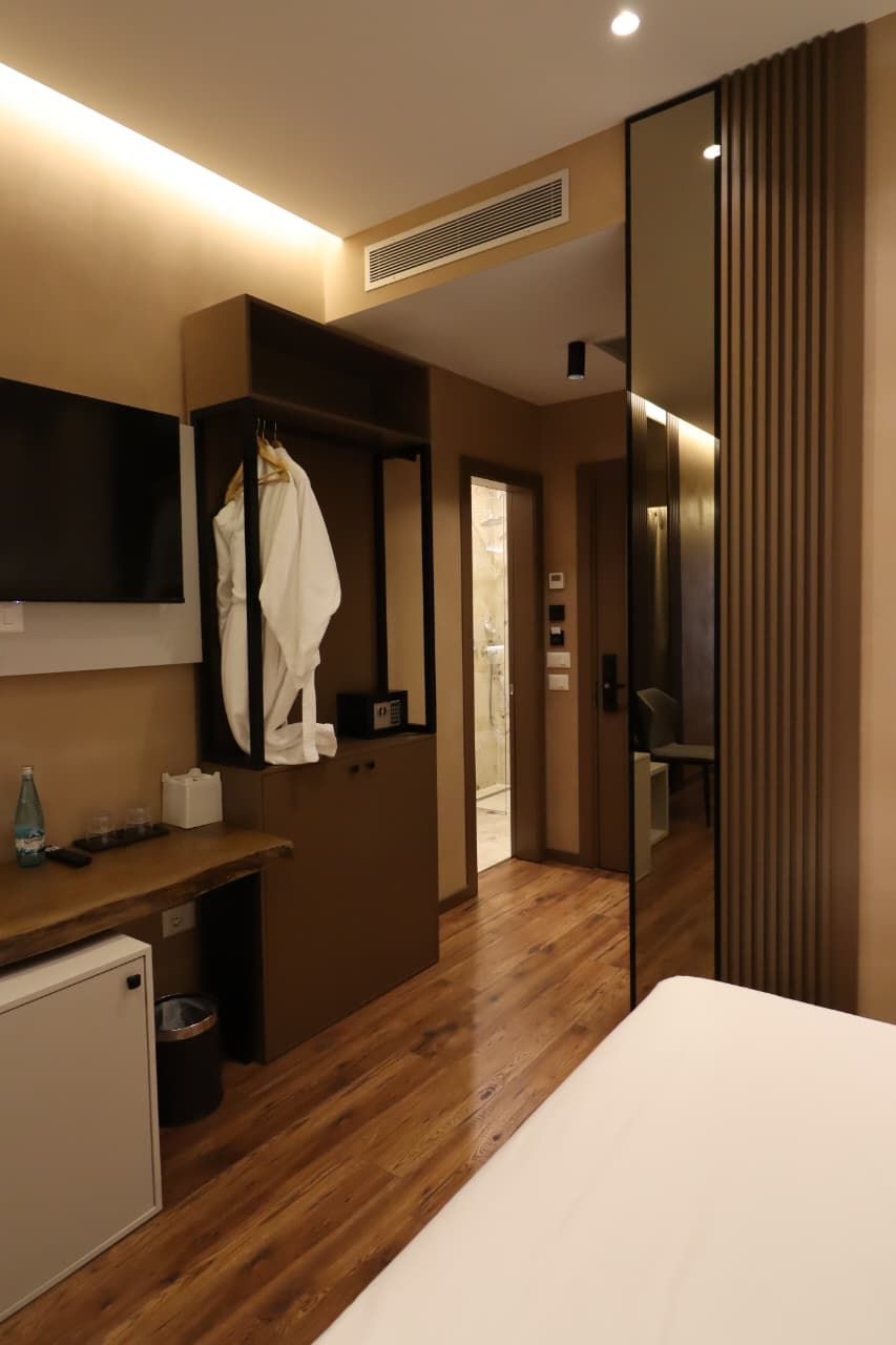 A hotel room with a wooden floor, a bed, a desk with a mirror, a TV, and a wardrobe.