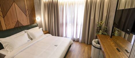 A modern hotel room with a large bed, a wooden headboard, a desk with a chair, a TV, and a window with curtains.