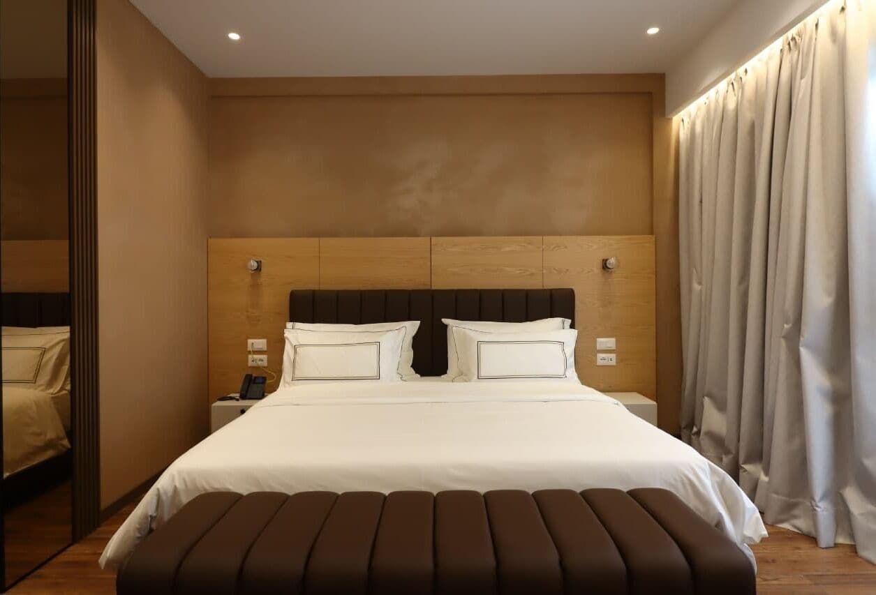 Een moderne hotelkamer met een groot bed, een houten hoofdbord, een nachtkastje met telefoon, en een groot raam met gordijnen.