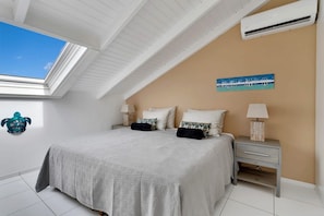 A bedroom with a skylight, a bed with a grey bedspread, two bedside tables with lamps, and a wall-mounted air conditioning unit. - Belmar Oceanfront Apartments 12 located directly on the sea with diving facilities (Kralendijk)