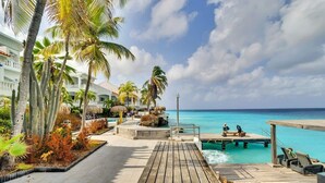 Eine Holzterrasse führt zu einem Pool mit Liegestühlen, umgeben von Palmen und einem Gebäude mit Balkon.