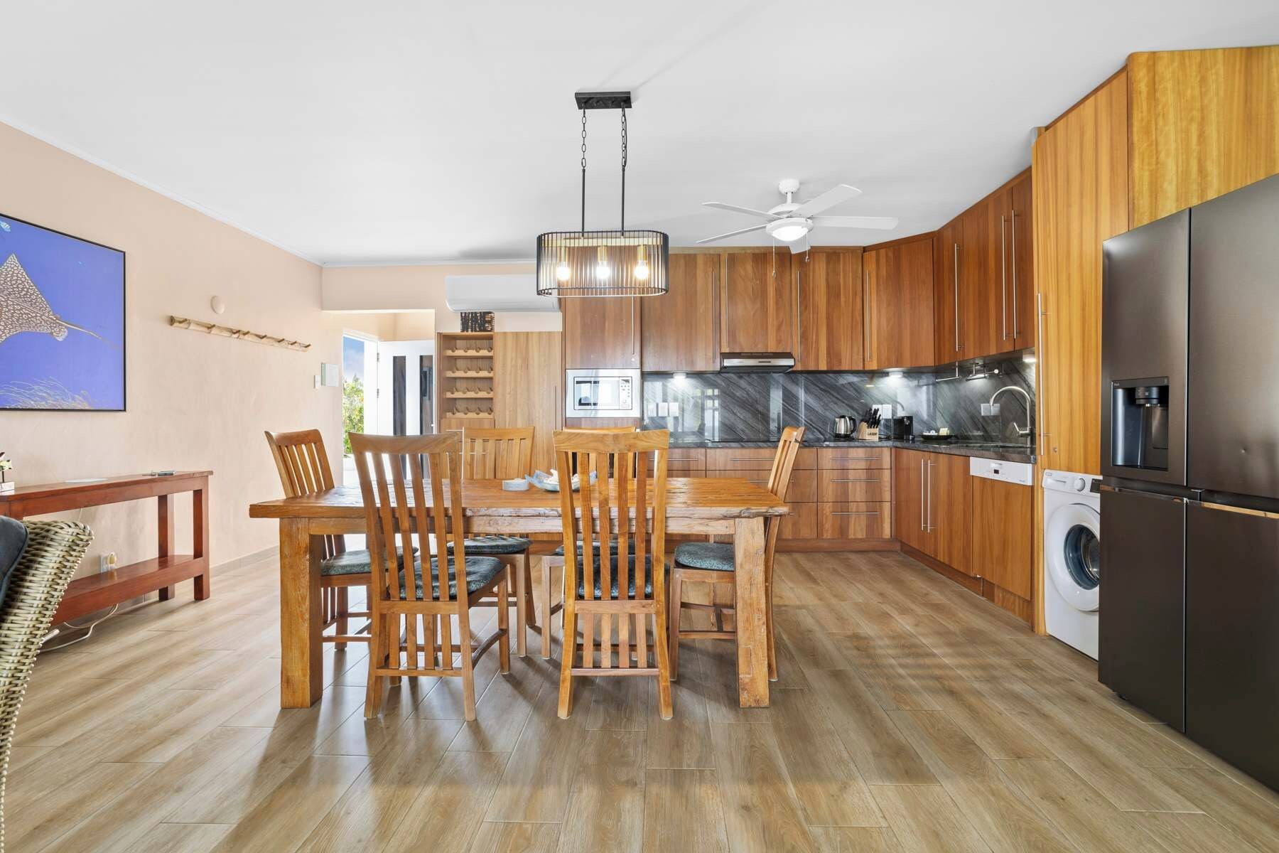 A modern kitchen with wooden cabinets, a dining area with a wooden table and chairs, a ceiling fan, and a refrigerator.