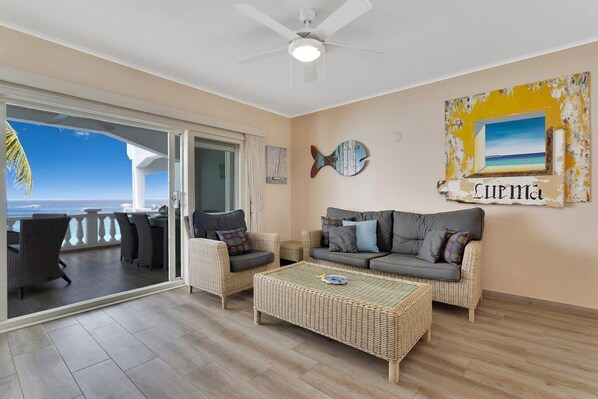 Un salon avec vue sur la mer, un ventilateur de plafond et un mur décoré d’un motif de poissons et d’un tableau encadré.