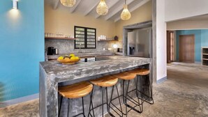 A modern kitchen with a central island, bar stools, hanging light fixtures, and a view of the adjacent living area. - Villa Heros Sabadeco (Kralendijk)
