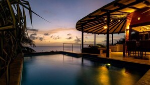 A poolside area with a view of the ocean at dusk, a covered seating area, and a bar. - Villa Kokopelli with stunning sea views and a refreshing private pool (Kralendijk)