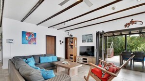 A modern living room with a sofa, coffee table, and a rocking chair. There is a television and a bookshelf. The room has a large window and a view of greenery outside. - Villa Kokopelli with stunning sea views and a refreshing private pool (Kralendijk)