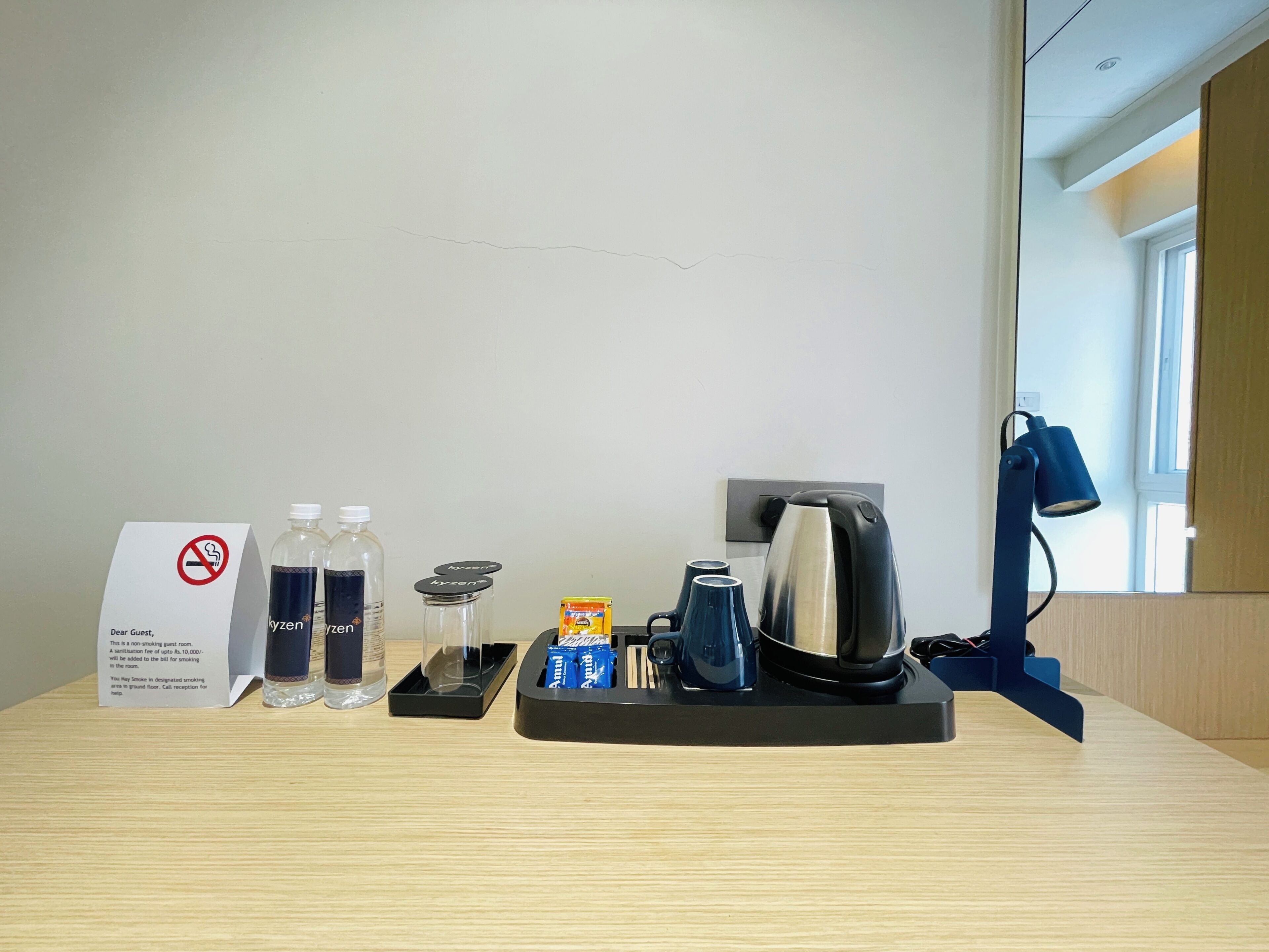 A hotel room with a no-smoking sign, a tray with toiletries, a kettle, and a desk lamp.