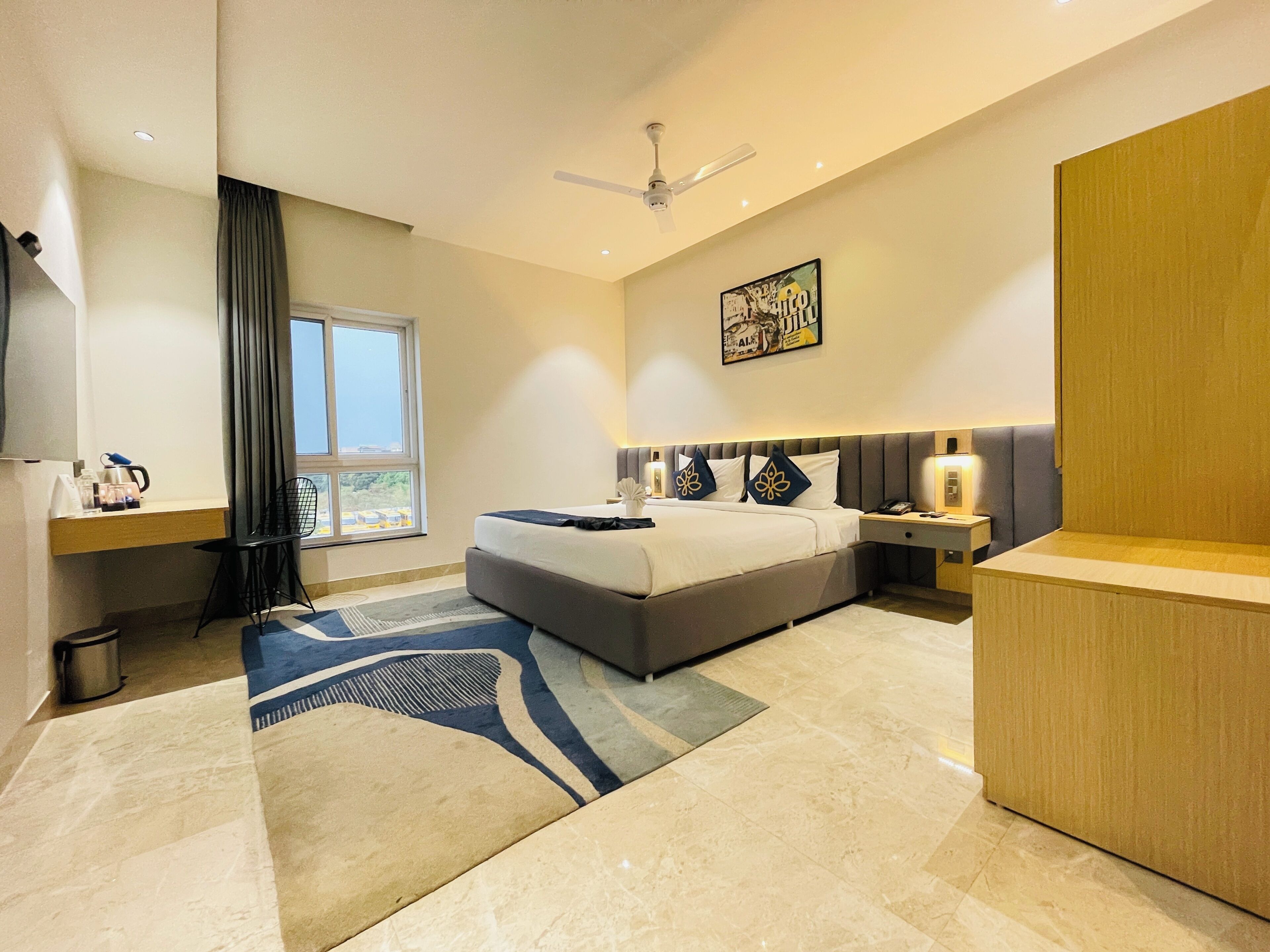 A modern hotel room with a large bed, a desk, a chair, a window, and a ceiling fan.