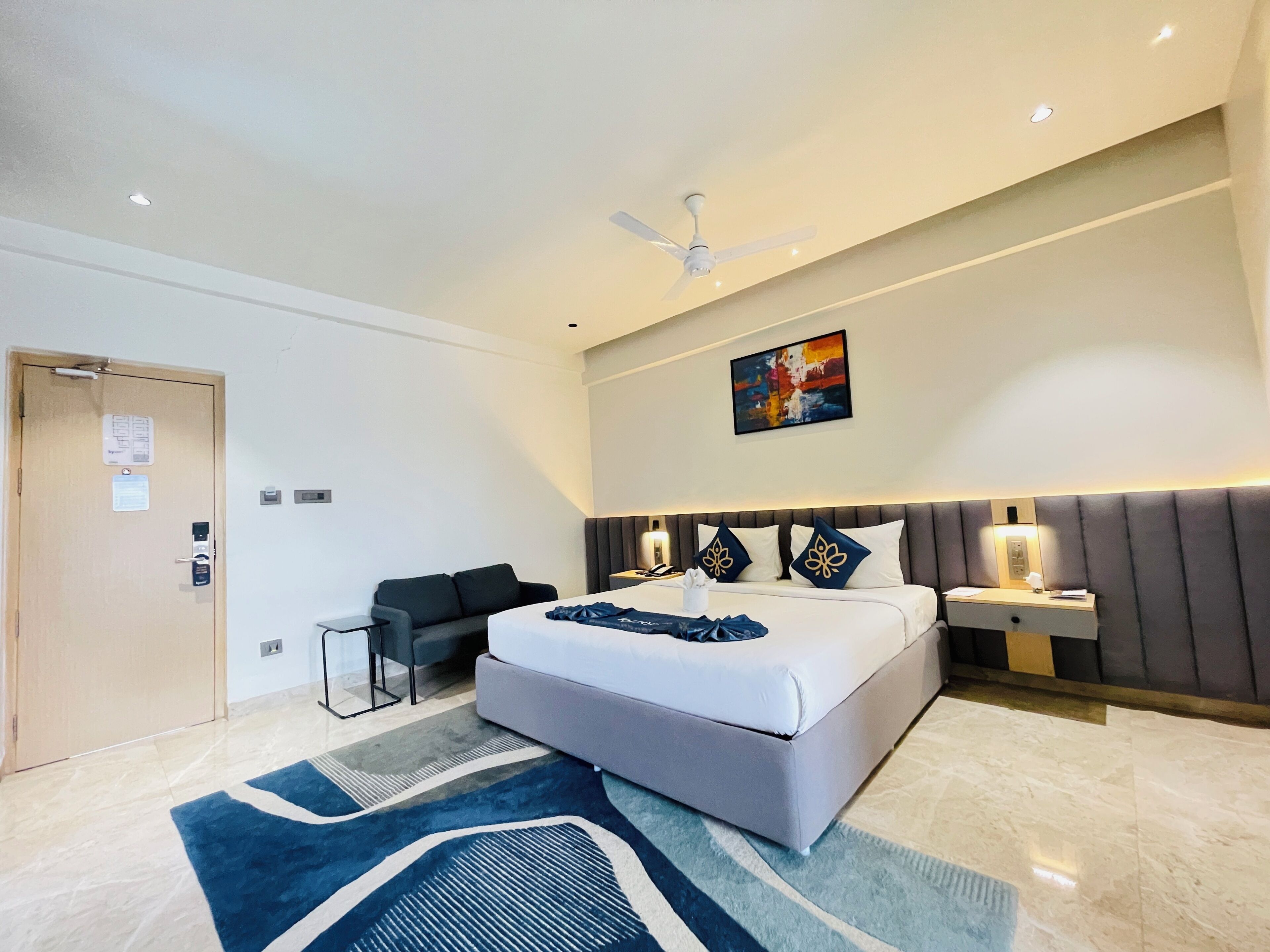 Une chambre d’hôtel moderne avec un grand lit, un ventilateur de plafond, un tableau, une table de chevet et un coin salon.