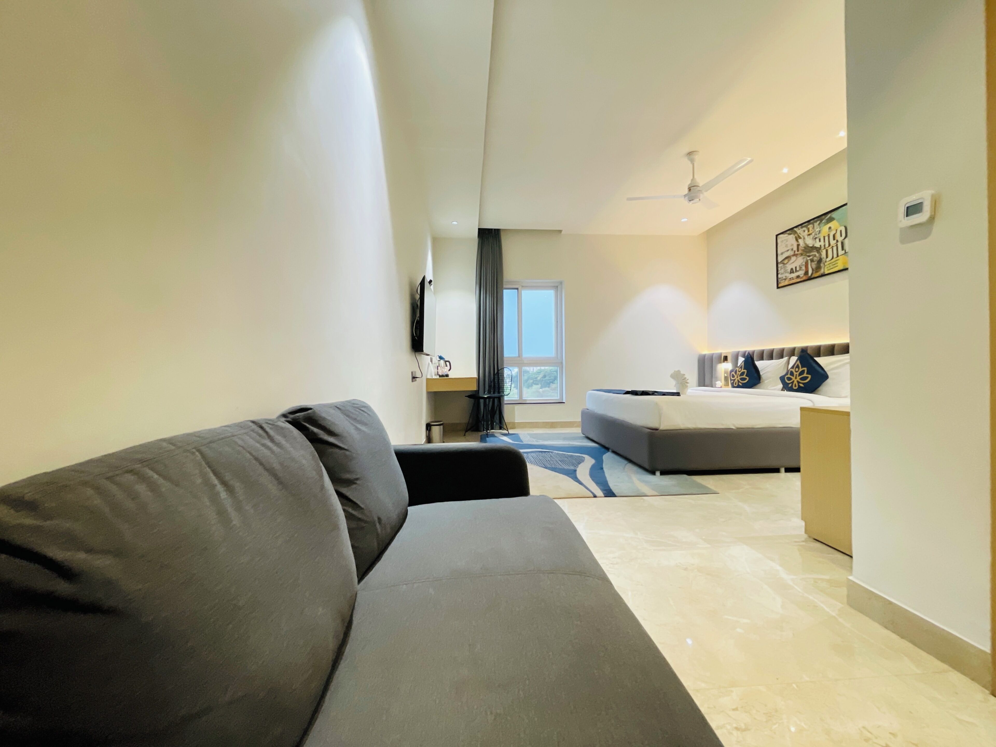 Une chambre d’hôtel moderne avec un canapé gris, un lit agrémenté d’oreillers décoratifs, un ventilateur de plafond et une grande fenêtre.