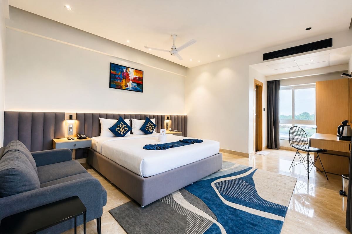 A modern hotel room with a large bed, a desk, a chair, a sofa, and a painting on the wall.