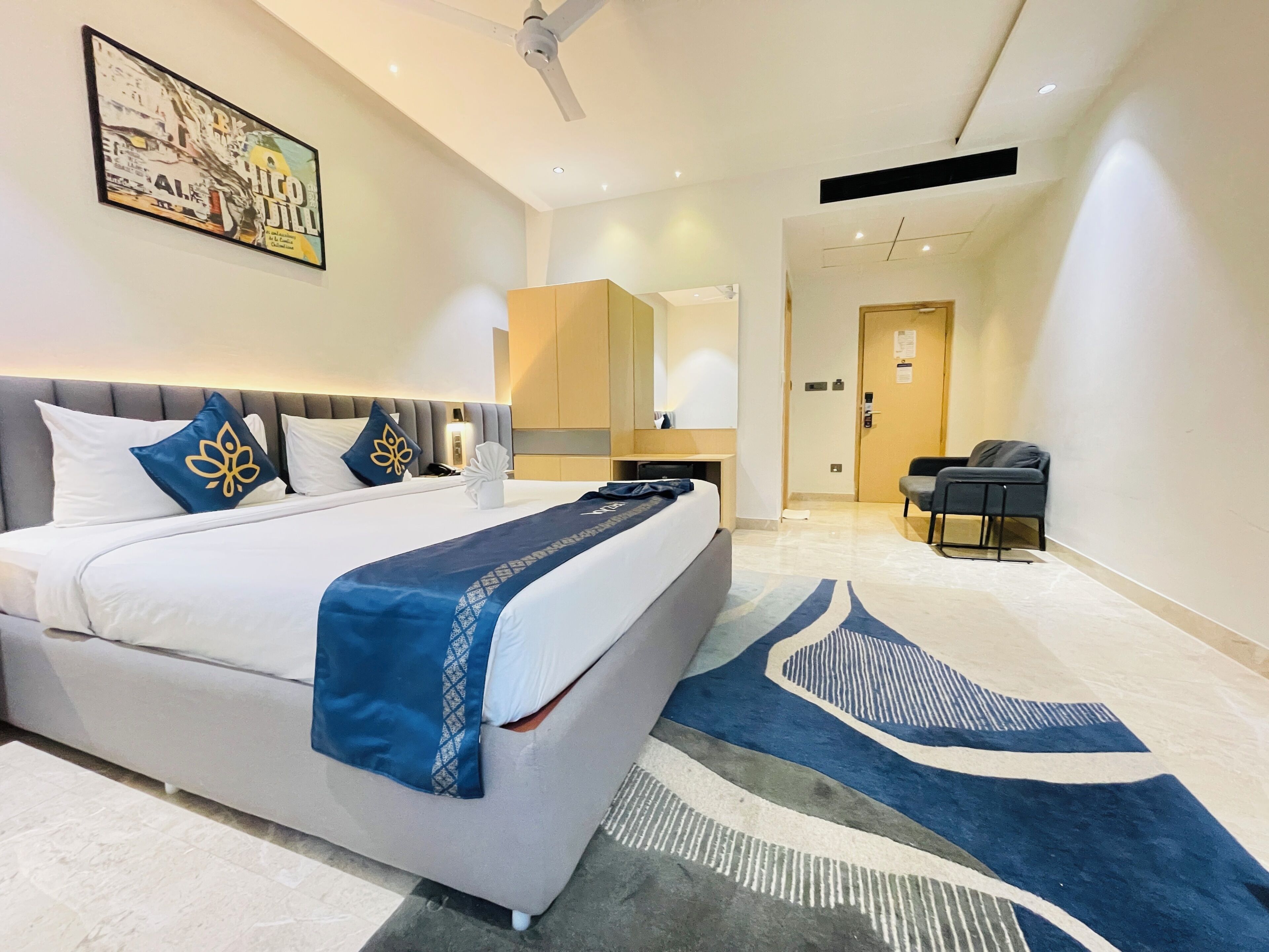 A hotel room with a bed, a desk, a chair, a ceiling fan, and a painting on the wall.