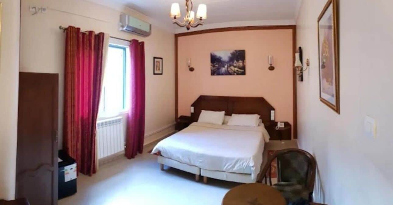 A bedroom with a bed, a chair, a radiator, a window with curtains, and a painting on the wall.