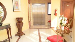 A lobby with a marble floor, a wooden chair with a red cushion, a small table, a mirror, and an elevator. - Ambassadeurs Oran (Oran)