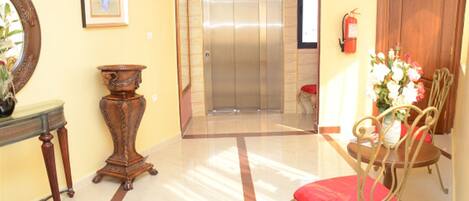 A lobby with a marble floor, a wooden chair with a red cushion, a small table, a mirror, and an elevator.