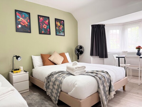 A neatly arranged bedroom with a bed, bedside tables, a desk, and a chair. There are three framed artworks on the wall, a fan, and a window with curtains. - Charming Studio 30min to Central London (Wembley)