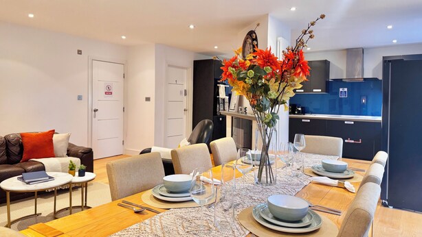 Un comedor moderno con una mesa larga preparada para seis personas, rodeada de sillas. Hay un florero con flores sobre la mesa, y la habitación tiene una paleta de colores neutros con paredes blancas y una cocina oscura al fondo.