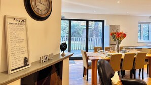Un comedor con una mesa de madera dispuesta para cuatro personas, un reloj en la pared y una lista enmarcada con las normas de la casa.