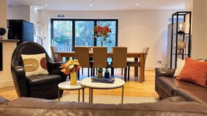A modern living room with a leather sofa, a dining table with chairs, a coffee table with a book and flowers, and a bookshelf with decorative items. - Family-Friendly En-suite 30min to Central London (Wembley)