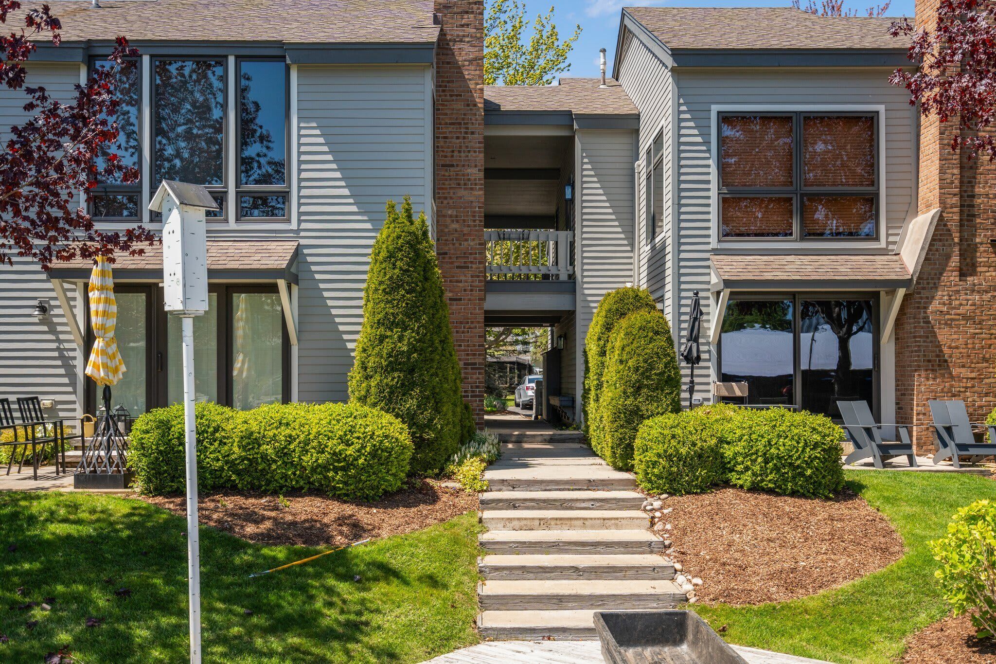 A two-story residential building with a front porch, a small garden with trimmed bushes, and a pathway leading to the entrance.